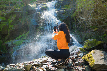 Woman standing in front of waterfall and taking pictures with the phone  - Powered by Adobe