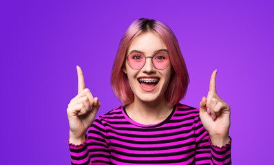 Excited pink woman in braces, opened mouth, wear sunglasses glasses spectacles advertise showing area for sales slogan text, isolated violet purple background. Dental care, ophthalmology ad concept.