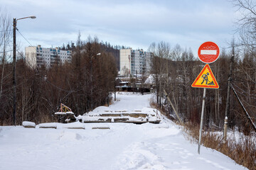 View of a snow-covered road and a collapsed bridge. Residential buildings in the distance. On the side of the road there are road signs “No Entry” and “Road Works”. Tynda, Amur region, Russia. - 794084624