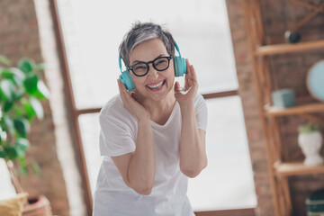 Photo of nice senior woman headphones listen music dressed white clothes cozy living room home house flat indoors pension concept