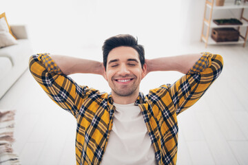 Photo of handsome cheerful guy dressed plaid shirt smiling arms behind head enjoying weekend indoors room home house
