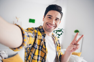 Photo of handsome cheerful guy dressed plaid shirt recording video showing v-sign indoors room home...