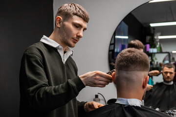 Focused barber cuts client hair on occiput with trimmer in barbershop. Male hairstylist does stylish hairdo to man in beauty salon. Haircut shape