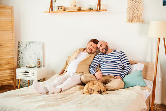 A gay couple, alongside their French Bulldog, lounging on a bed.