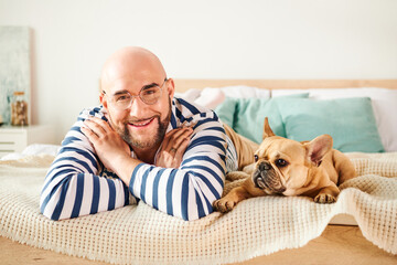 Handsome man with glasses relaxing on bed next to his loyal French Bulldog.
