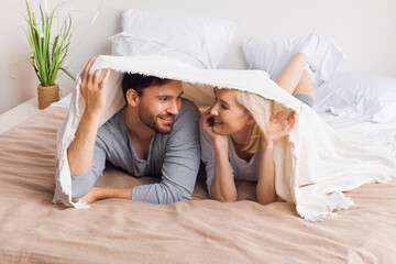 Portrait image - young inlove couple look at each other, covered with sheet-blanket over heads, lying lay on bed. Woman, brunette bearded man at home. Love, relationship, happy family, dating.