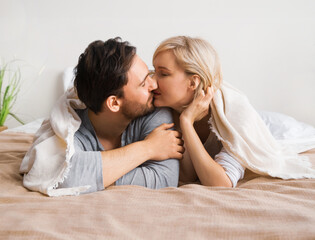 Portrait image - inlove kissing couple with closed eyes, covered with sheet-blanket over heads, lying lay on bed. Woman, brunette bearded man at home. Love, relationship, happy family, dating.