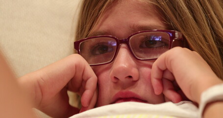 Closeup of little girl face watching scary movie screen child wearing glasses paying attention to...