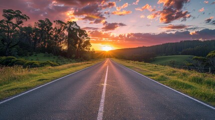 A road with a sunset in the background. The sky is orange and the sun is setting. The road is empty and there are no cars on it
