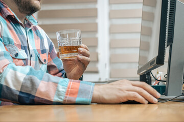 Drunk and frustrated man in the office while working with pc, sitting at the work table with glass of alcohol drink, whiskey or brandy, alcoholism and bad habits concept