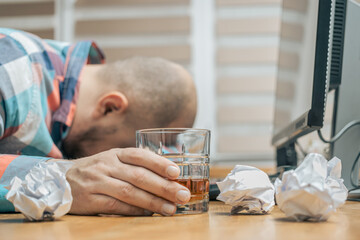 Drunk and frustrated man in the office while working with pc, sitting at the work table with glass of alcohol drink, whiskey or brandy, alcoholism and bad habits concept