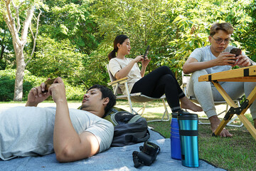 Group of three asian friends carrying backpacks, woman and man sitting on picnic and young man sleeping on camping mat, picnicking in the forest, everyone holding smartphones playing games