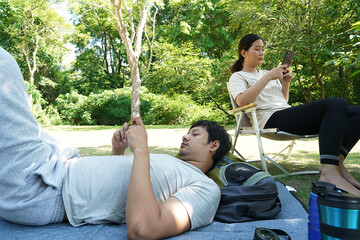 Asian couple, girlfriend sitting on picnic, young man sleeping on a camping mat, picnicking in the forest, both holding smartphones playing games with social network in the forest.