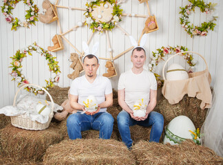 Two man with rabbit ears sitting on the haystack and holding Easter eggs