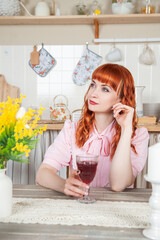 Beautiful redhaired woman with glass of wine sitting in the kitchen at home