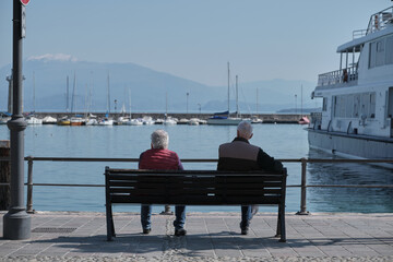 Rear view of women and men relaxing on a bench overlooking the boat station, mountains, water.