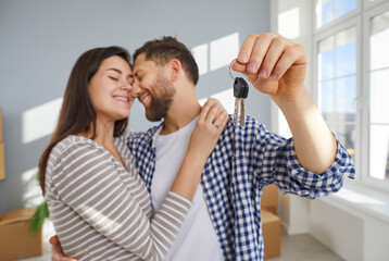 Happy young couple holding a key in hands standing in the living room at home hugging enjoying real estate or car purchase, smiling and celebrating moving day. Relocating, mortgage concept.