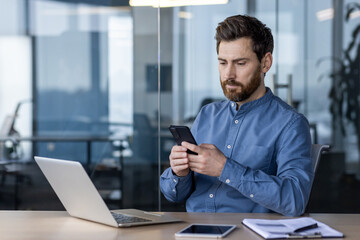 Obraz premium A serious young man sits in the office at the desk and uses a mobile phone in his hands