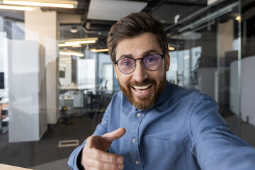 Close-up portrait of a young smiling man in glasses and a shirt standing in a modern office and chatting online while pointing his finger at the camera