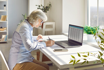 Concentrated serious senior accountant woman sitting on workplace at the desk working on laptop...