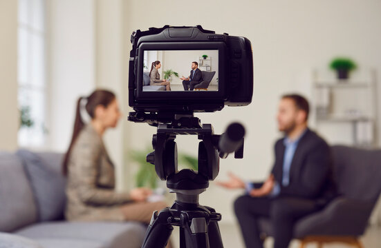 Camera on tripod foreground, businessman giving interview to journalist in office, female reporter interviewing guest speaker for podcast, radio, business talk, media show, broadcasting success story