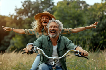 In a daylight scene, a happy middle-aged couple rides a bike through a rural area. The woman, with blonde curly hair and a hat, smiles with arms outstretched in joy. The man, sporting a beard and glas