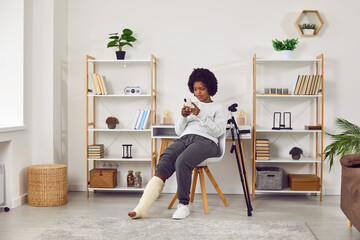 Young African American woman with a broken leg looking at her cell phone with a surprised face expression while sitting on a chair with crutches in the living room at home