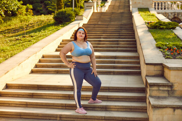 Happy young fat woman in sportswear looking at the camera standing confidently on the stairs in the summer city park ready for sport fit exercises. Workout in nature and fitness outdoors concept.