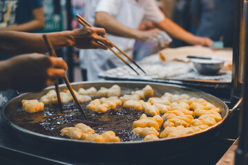 A vendor skillfully uses chopsticks to fry dough in a large pan at a bustling street food market.