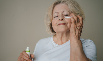 Close-up of senior gray hair woman applying anti-aging cream under the eyes against grey background. Skin treatment. Makeup product.
