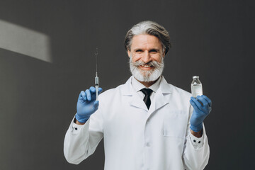 Gray-haired senior doctor in white coat holding syringe and bottle with injection, isolated on gray background.