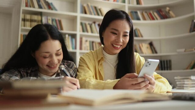 Charming students girls interracial classmates holding smartphone and show funny pictures at university library after lessons Multiethnic friends at university