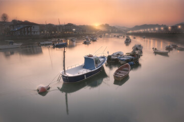 Boats at dawn in the Plentzia estuary with the sun rising behind the mountains leaving a warm...