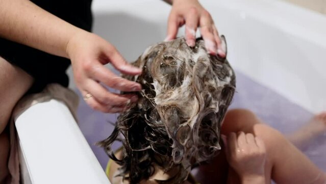 A mother washes her daughter's hair in the shower. A woman lathers shampoo on a child's head. The girl takes a bath. Water procedures. Baby hair care. Children's hygiene. High quality 4k video
