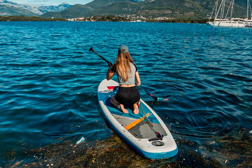 Adventurous Woman Paddleboarding on Crystal Blue Water with Mountainous Landscape and Sailboats in the Background
