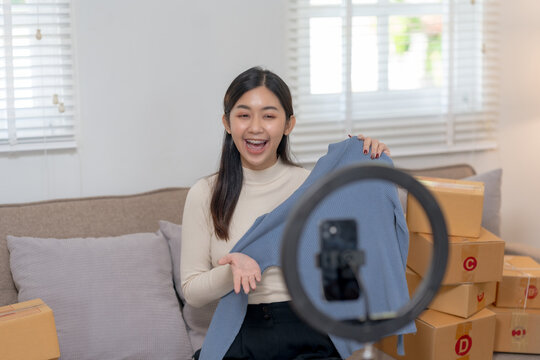 Smiling woman showcasing a blue sweater for an online audience with a smartphone and ring light. Online selling and social media marketing concept.