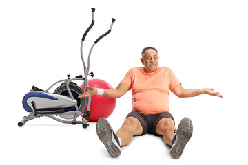 Mature man in sportswear sitting on the ground next to a stationary bike and fitness ball