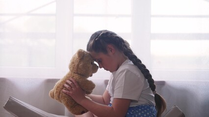 depressed girl child sitting by the window with her favorite toy teddy bear upset. problem of...