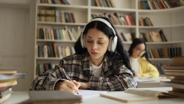 Pretty young woman student with headphones listening to music while writes notes for essay study for class preparing for exams test make research for project using books at university library
