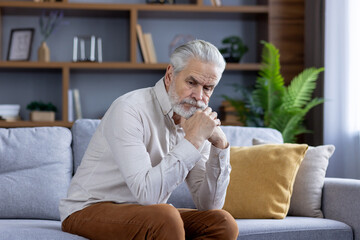 A lonely senior gray-haired man sits upset at home on the sofa, and pensively contemplates, resting...