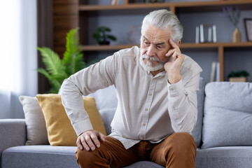 Upset and tired senior gray-haired man sitting on the sofa at home and leaning his head on his hand...