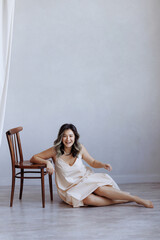 A woman is sitting on a wooden chair in a room with white walls. She is wearing a dress and smiling. Concept of relaxation and comfort, as the woman is at ease in her surroundings