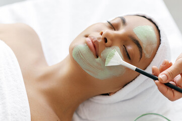 Facial treatments, wrinkle prevention, facial cleansing. Therapist applying green face mask on the face of a beautiful brazilian or hispanic young woman. Skincare, beauty procedure concept
