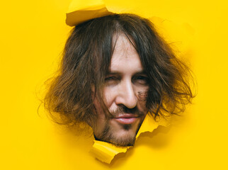 A long-haired, shaggy man with a beard and mustache with a smiling expression on his face looks out from a torn hole in yellow paper. Concept of originality and old-fashioned people. Hippie.