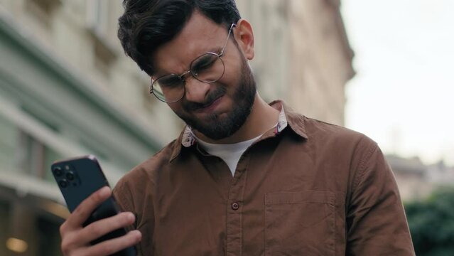 Close up upset unhappy Indian Arabian ethnic man young guy businessman male using looking mobile phone bad connection losing fortune loss fail touch head why negative result outdoors city urban street