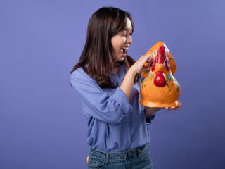 Excited asian woman holding a colorful rooster piggy bank, with a coin in hand, against a purple...