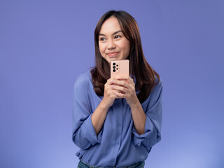 Naklejka premium Excited Asian woman celebrating good news on her smartphone, wearing a stylish blue blouse and jeans, isolated on a purple background