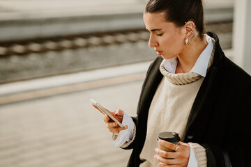 Side view of a fashionable woman standing at train station with phone.