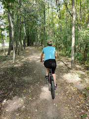 A young blonde woman rides a bicycle in the park on a path. Rear view. Tourism, sports and outdoor recreation