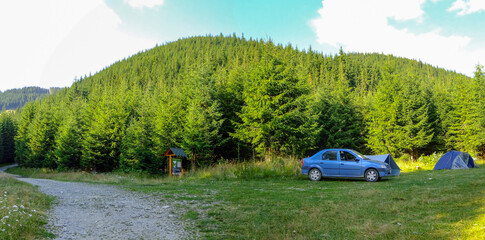 A peaceful forest campsite showcases a blue car parked beside a tent, surrounded by tall green trees under a clear blue sky on a sunny afternoon. Lotru Mountains, Carpathia, Romania.